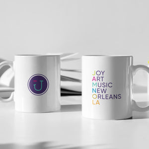 2 White Coffee Cups on a white table, the left one displays a JAMNOLA wink face logo, on the right mug are the words Joy Art Music New Orleans.