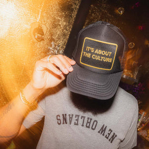 Person wearing a black cap with 'It's About the Culture' text and a gray 'New Orleans' shirt, against a blurred background.
