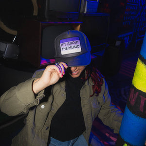Person wearing black trucker hat with purple ombre It's About the Music applique.