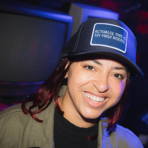Person wearing a blue trucker hat that says Actually this is my first rodeo smiling in a dimly lit room.