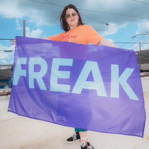 Person holding a purple flag with 'FREAK' printed on it against a blue sky.