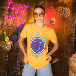 Person wearing a marigold t-shirt with a JAMNOLA logo in front of a neon 'FEAST MODE' sign.