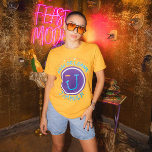Person wearing a yellow JAMNOLA t-shirt with logo and text, standing in front of a gold wall with neon signs.