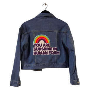 Blue denim jacket with embroidered text and rainbow design on a white surface.