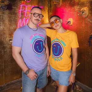 Two people wearing Hugo Gyrl's t-shirts with a JAMNOLA wink face design in front of a neon sign.