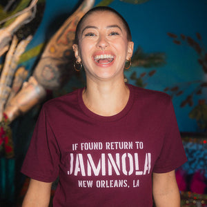 Person wearing a maroon t-shirt that says 'If found return to JAMNOLA', standing in front of a colorful background.