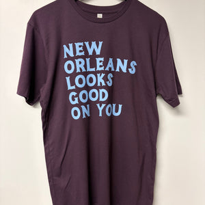 Purple t-shirt with text 'New Orleans Looks Good On You' on a beige background