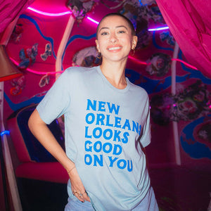 Person wearing a light blue t-shirt with 'New Orleans Looks Good On You' text in a colorful indoor setting.