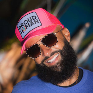 Man wearing a pink trucker hat with 'PROUD HUMAN' text and sunglasses, outdoors.