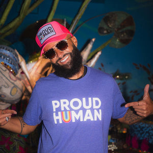 Person wearing a blue 'PROUD HUMAN' t-shirt and pink cap with sunglasses, standing in front of decorative plants.