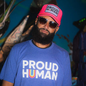 Man wearing a blue 'PROUD HUMAN' t-shirt and pink cap with sunglasses, against a colorful background.