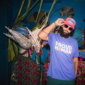 Person wearing a 'Proud Human' shirt and pink trucker hat, standing in front of colorful decorations.