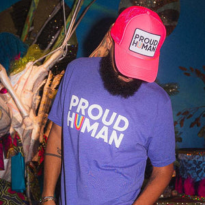 Person wearing a purple 'PROUD HUMAN' t-shirt and matching hot pink cap with text, standing against a colorful background.
