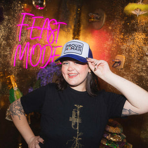 Woman posing in a gold room wearing a white trucker hat with purple bill that says Proud Human, and a black t-shirt with a trumpet.