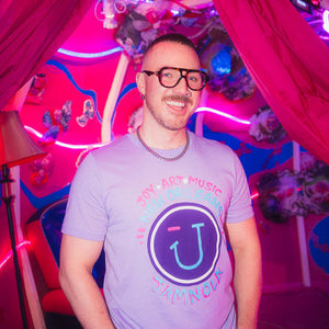 Man wearing a lavender JAMNOLA t-shirt with a logo and text in a colorful, neon-lit room.