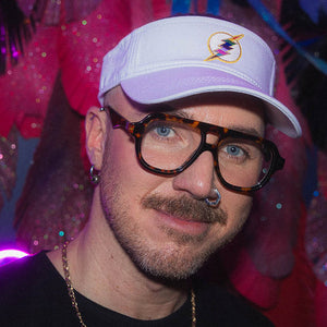 Person wearing a white sun visor with a colorful thunderbolt and large glasses, against a vibrant background.