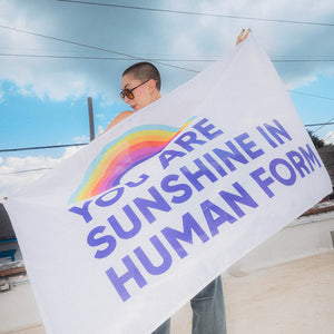 Person holding a You are Sunshine in Human Form flag against a blue sky.