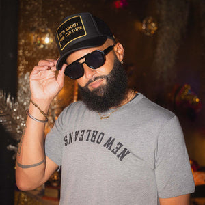 Man wearing a black hat and sunglasses and a gray tee-shirt with New Orleans written upside down, with a blurred background