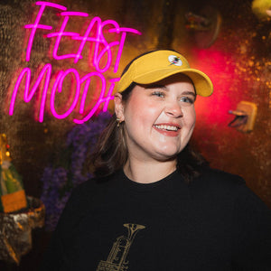 Person wearing a yellow sunvisor with a rainbow thunderbolt, smiling in front of a neon sign and colorful background.