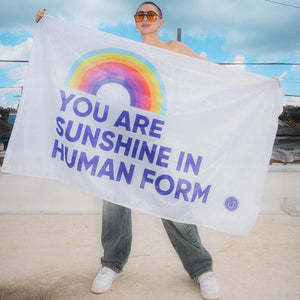 Person holding a You are Sunshine in Human Form flag with a rainbow against a blue sky.