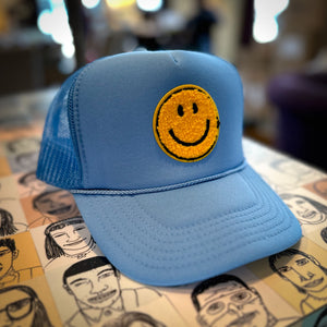 A powder blue trucker hat with a yellow smiley face sitting on a table. 