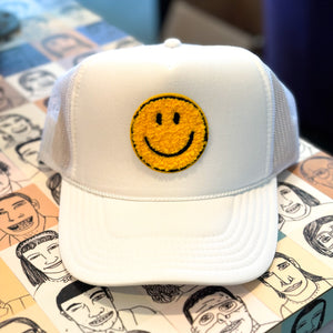 A white trucker hat with a textured yellow happy face on a table.